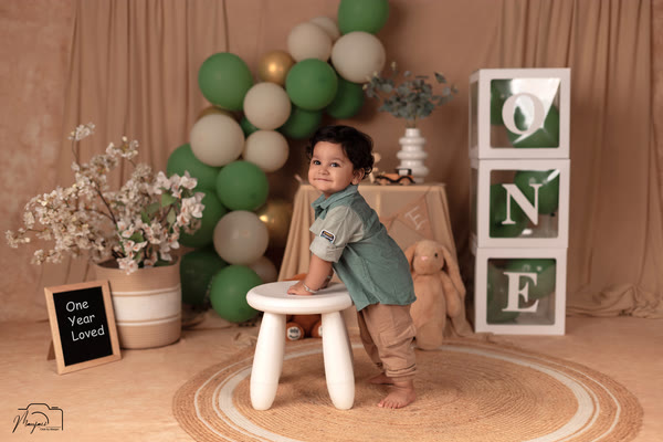 Toddler standing at a stool beside green balloons and ONE blocks