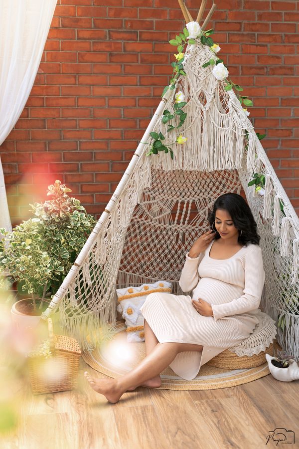 Expectant mother in a cream knit dress seated beneath a macrame teepee