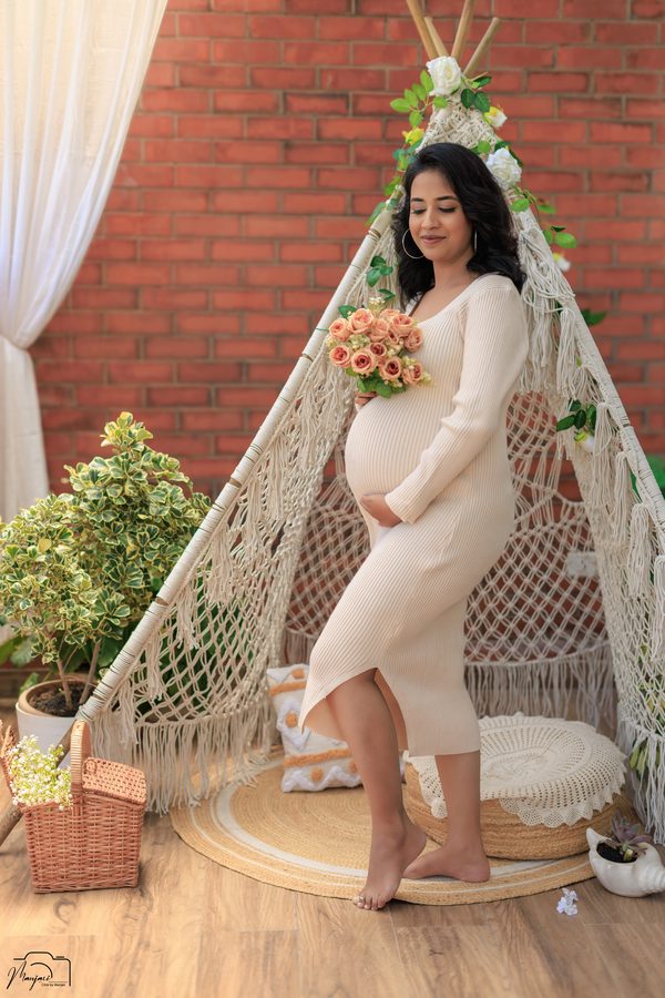 Expectant mother in a cream dress holding a bouquet beside a macrame teepee
