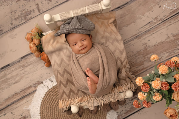 Smiling newborn in a grey cap on a miniature wooden bed with orange flowers